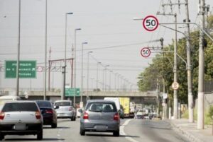 Uma imagem de carros no trânsito. Ao alto, placas definem o limite de velocidade em 50 km/h