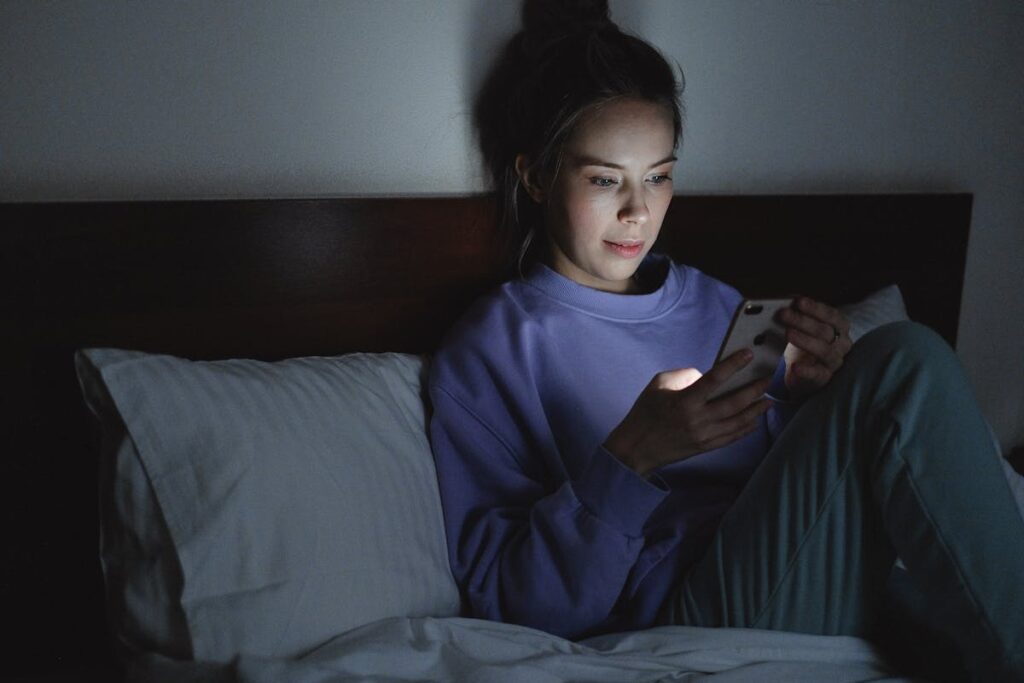 Jovem sentada na cama com a luz do quarto apagada olha para a tela do celular